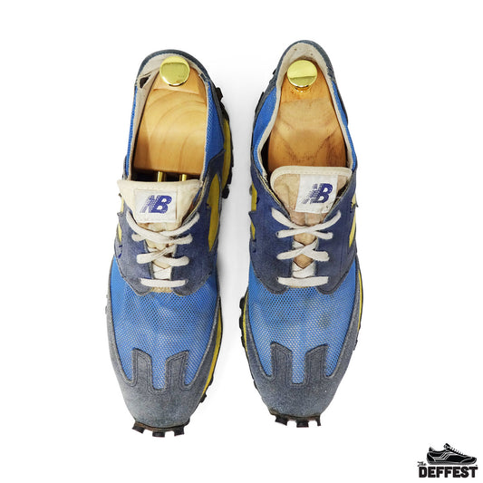 A pair of vintage New Balance 355 sneakers shown from a top-down view, featuring a blue and gray suede/mesh mix with yellow accents, white laces, and wooden shoe trees inside.