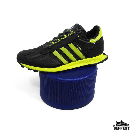A black Adidas Formel 1 running shoe with neon yellow-green accents, displayed on a blue cylindrical bottle cap riser against a white background.
