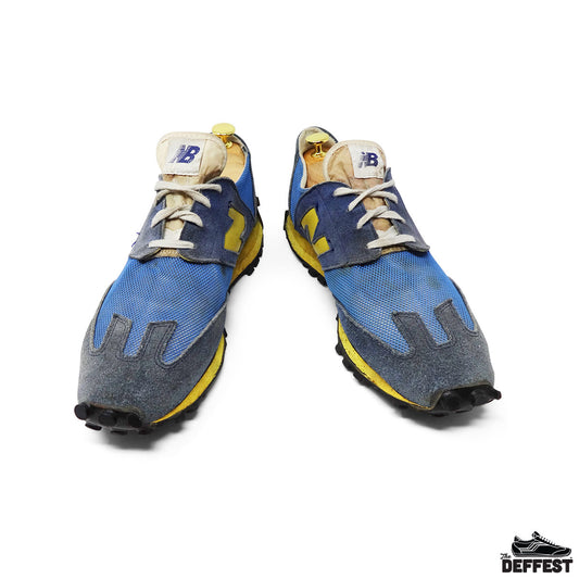 A pair of New Balance 355 sneakers in a washed blue, yellow, and gray colorway, shown from a front-facing perspective on a white background with a 'DEFFEST' brand watermark in the bottom right corner.